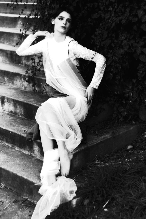 Model in a sheer white dress reclining on old stone steps beside ivy, black and white